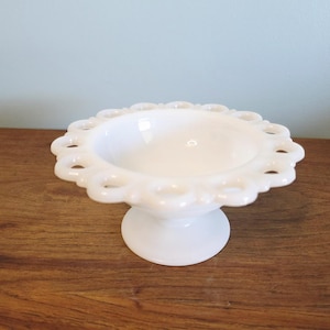 Puede incluir: Una taza de cerámica blanca con asa negra y el texto "But First, Coffee" en negrita negra. La taza se encuentra sobre un fondo borroso, sugiriendo un ambiente acogedor.