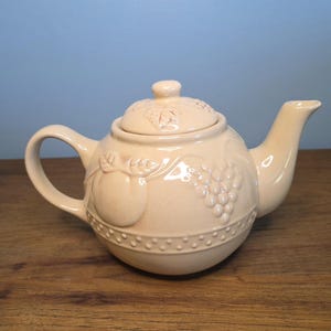 Pode incluir: Bule de chá de cerâmica creme com tampa e alça. O bule apresenta desenhos em relevo de frutas e uvas, com uma borda decorativa ao redor da base. O bico é curvo e a tampa tem um pequeno botão. O bule está sobre uma superfície de madeira.