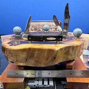 May include: A Case knife with three blades displayed on a wooden base. The knife has a black handle and silver blades. The base is a natural wood slab with a dark finish. Two small, gray spheres are on the base. A ruler shows cm.