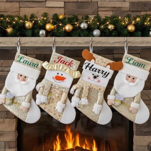 Peut inclure: Quatre bas de Noël dorés suspendus au-dessus d'une cheminée. Chaque bas représente un personnage différent : le Père Noël, un bonhomme de neige, un renne et le Père Noël. Chaque bas porte un nom brodé : Laura, Jack, Harry et Liam.