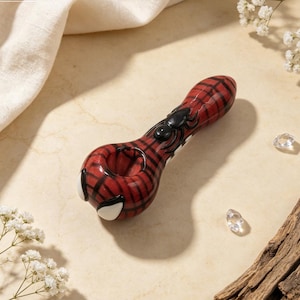 May include: A red and black glass pipe featuring a spider web pattern, a raised black spider figure on the stem, and two white eye-shaped accents on the bowl. The pipe has a smooth, glossy finish and a rounded shape with a central opening.