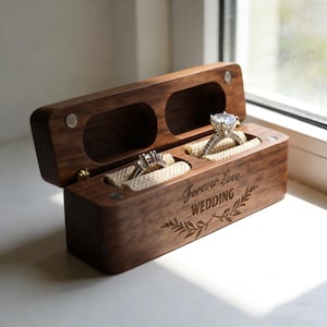 May include: A wooden ring box with two compartments, each holding a ring. The box is engraved with "Forever Love WEDDING" and a leaf design. The rings have silver bands and clear stones. The box is open, revealing the rings.