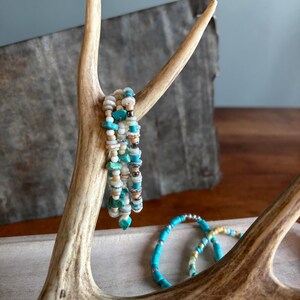 May include: A display of beaded bracelets in shades of turquoise, white, and tan. The bracelets are arranged on a natural antler, with a wooden tray in the background. The bracelets feature various bead sizes and textures.