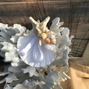 May include: White coral with a white seashell and a white satin ribbon. The seashell is decorated with small seashells in shades of white, yellow, and brown.