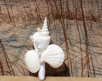 May include: A white seashell bouquet with pearls and a white ribbon. The bouquet is on a sandy background.
