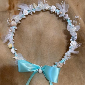 May include: A handcrafted circlet, featuring a light blue ribbon base adorned with seashells, white flowers, pearls, and sheer white accents. A matching light blue satin bow is centered at the front, creating a delicate, ocean-themed accessory.