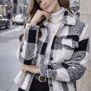 May include: A black and white checkered fleece jacket with a button-up front. The jacket is worn over a white turtleneck and paired with black jeans. A gold belt and jewelry complete the look. The background shows a city street.