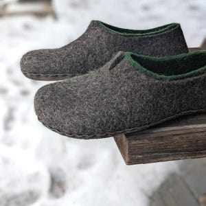 May include: A pair of gray felted wool slippers with a dark green trim. The slippers are sitting on a wooden surface, with a snowy background. The slippers appear to be handmade.
