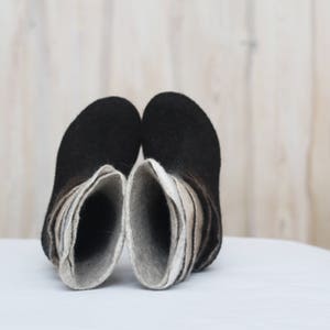 Pode incluir: Um par de botas de feltro preto com um design em camadas em tons de cinza e branco. As botas estão viradas do avesso, mostrando as camadas de feltro.