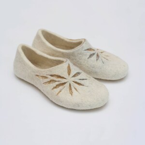 May include: A pair of off-white felt slippers. Each slipper features a brown starburst design on the top. The slippers have a rounded toe and a low profile, suggesting a comfortable, indoor shoe.