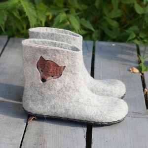 May include: A pair of gray felt slippers with a fox design on the front. The slippers are on a wooden surface.