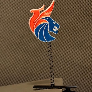 May include: A decorative clip featuring a lion's head design with orange, blue, and white colors. The lion's head is attached to a black spring and clip base. The clip is designed to hold notes or photos.