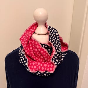 Op de afbeelding: Een omkeerbare infinity sjaal met een roze en witte stippenpatroon aan de ene kant en een marineblauw en wit stippenpatroon aan de andere kant. De sjaal is over een mannequin gedrapeerd.