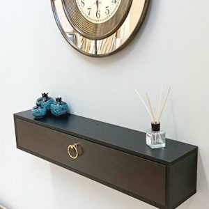 Puede incluir: Una estantería flotante de pared marrón oscuro con un tirador de anillo dorado. La estantería contiene pequeños objetos decorativos azules y un frasco difusor de vidrio. Un gran reloj redondo con marco dorado cuelga encima.