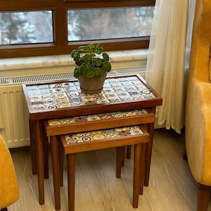 Può includere: Set di tre tavolini a incastro con struttura in legno e piani a mosaico. I tavoli sono di diverse dimensioni, il più grande misura circa 61 cm di larghezza, 46 cm di profondità e 51 cm di altezza. Una piccola pianta in vaso è sul tavolo superiore.