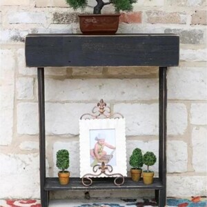 Puede incluir: Una estantería de madera negra con un pequeño bonsái en una maceta marrón en la parte superior. La balda inferior contiene un marco de fotos blanco con un soporte de metal decorativo y dos pequeños árboles topiarios en macetas.