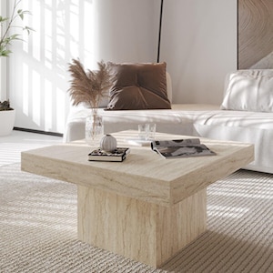 Puede incluir: Una mesa de centro cuadrada de color beige claro con una superficie texturizada y una base sólida. La mesa está en un salón moderno, con un sofá, objetos decorativos y luz natural.