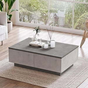 Puede incluir: Una mesa de centro rectangular moderna con una tapa gris efecto hormigón y laterales grises claros. La mesa tiene un perfil bajo y descansa sobre una base gris oscuro. Velas y una pequeña planta están dispuestas sobre la mesa.