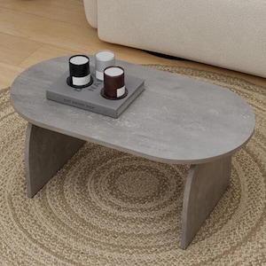 Puede incluir: Una mesa de centro ovalada gris con una superficie texturizada. La mesa tiene dos patas curvas y un libro con el texto "EAT DRINK NAP" encima. Tres pequeñas velas negras, blancas y marrones están sobre el libro. La mesa está sobre una alfombra redonda de yute.