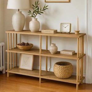 Può includere: Una consolle in legno chiaro con tre ripiani. I ripiani espongono vari oggetti decorativi, tra cui una lampada, vasi, un orologio, una candela, una ciotola di legno, un quadro incorniciato e un cestino intrecciato. Il tavolo ha supporti verticali.