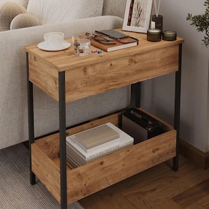 Puede incluir: Mesa auxiliar de madera de dos niveles con acabado de vetas de madera natural y estructura de metal negro. La balda superior contiene una taza y un platillo, un vaso y un cuaderno. La balda inferior contiene libros y un pequeño altavoz.