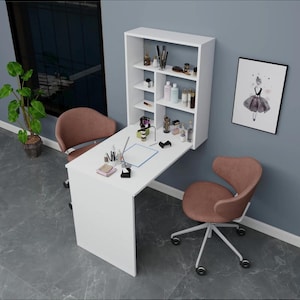 Puede incluir: Una estación de manicura blanca montada en la pared con una mesa plegable y estanterías empotradas. La estación incluye un escritorio, dos sillas giratorias marrones y una imagen enmarcada. Varios productos y herramientas de belleza se exhiben en los estantes y la mesa.