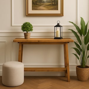Op de afbeelding: Een houten consoletafel met een onderste plank en schuine poten. Een kleine plant in een pot, een zwarte lantaarn met een brandende kaars en een decoratieve voetenbank zijn op of in de buurt van de tafel geplaatst. Een ingelijste landschapsschilderij hangt boven de tafel.