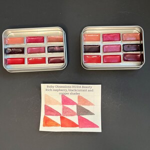 May include: Two silver rectangular palettes each containing 10 lip colors in shades of red, pink, and brown. A small card displays swatches of the same colors, with text that reads "Ruby Obsessions HUDA Beauty Rich raspberry, blackcurrant and copper shades."