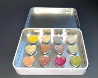 May include: A white rectangular tin with a silver interior, containing twelve heart-shaped items in various colours. The hearts are yellow, tan, grey, beige, gold, brown, glittery silver, green, orange, red-brown, pink, and green.