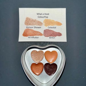 May include: A heart-shaped eyeshadow palette with four shades of eyeshadow in a silver container. The shades include shimmering gold, orange, copper, and deep brown. A card with the text "What a Hoot ColourPop" and the names of the shades is also present.