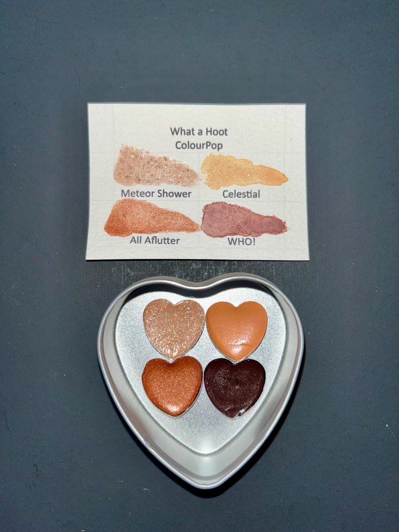 May include: A heart-shaped eyeshadow palette with four shades of eyeshadow in a silver container. The shades include shimmering gold, orange, copper, and deep brown. A card with the text "What a Hoot ColourPop" and the names of the shades is also present.