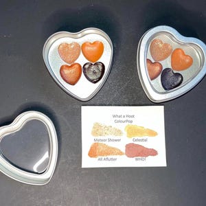 May include: Heart-shaped tins containing heart-shaped makeup in shades of bronze, orange, and black. A small card displays eyeshadow color swatches with the text "What a Hoot ColourPop". One empty heart-shaped tin is also present.