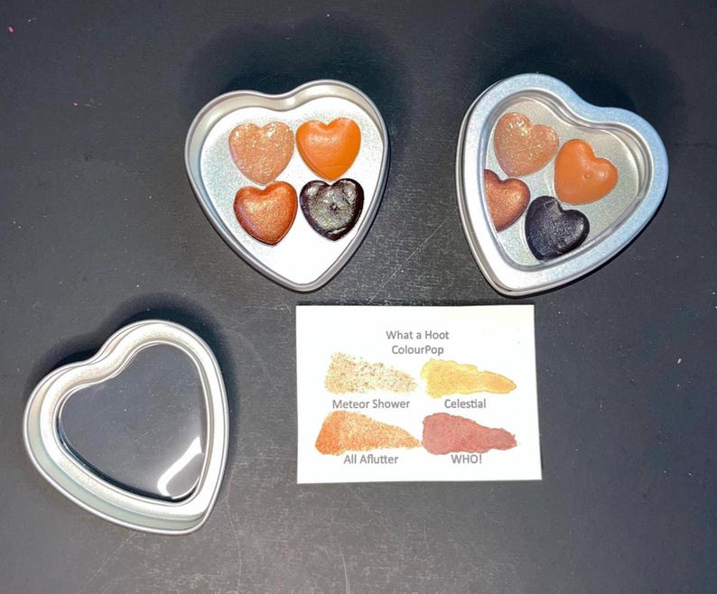 May include: Heart-shaped tins containing heart-shaped makeup in shades of bronze, orange, and black. A small card displays eyeshadow color swatches with the text "What a Hoot ColourPop". One empty heart-shaped tin is also present.