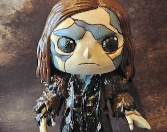 May include: A handcrafted figurine of a character with a distressed appearance. The figure has a cracked face with blue and cream tones, large black eyes, and brown hair. It wears a black coat and stands on a small, round base.