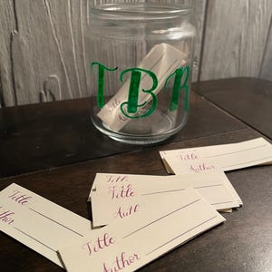 Pode incluir: Um frasco de vidro transparente com as letras TBR impressas em verde na parte frontal. Dentro do frasco e espalhadas à frente, encontram-se várias tiras de papel branco retangulares. Cada tira apresenta as palavras Title e Author impressas.