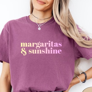May include: Berry-colored t-shirt with the text "margaritas & sunshine" in a gradient yellow and pink font. The shirt is a casual style, suitable for warm weather or a relaxed vibe.