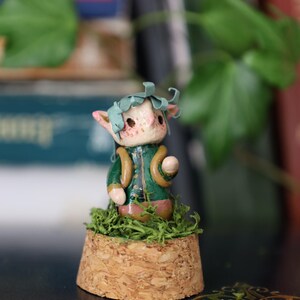 May include: A whimsical figurine of a small creature with pointed ears, wearing a green jacket and a leafy headpiece. The figure is set on a cork base covered in green moss. The overall aesthetic is fantasy-themed.