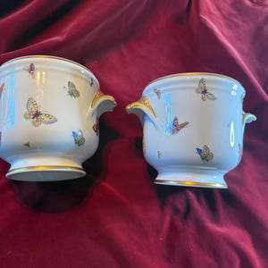 May include: Two white porcelain planters with gold trim and handles. The planters are decorated with colorful butterflies. The planters are on a red velvet surface.