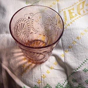 Puede incluir: Un vaso te&ntilde;ido de rosa con un dise&ntilde;o floral en relieve. El vaso est&aacute; sobre un pa&ntilde;o blanco con detalles bordados en amarillo y verde. El vaso tiene una base gruesa y un borde ancho.