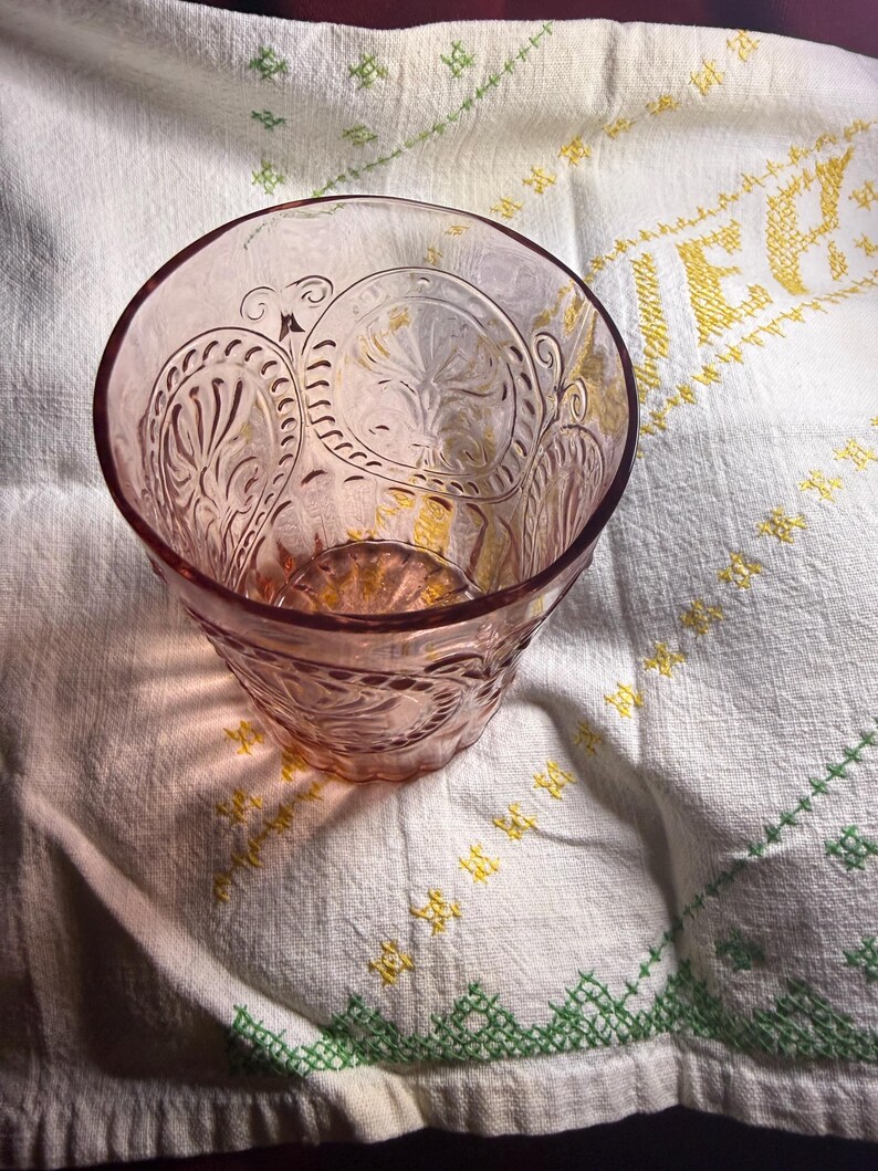 Puede incluir: Un vaso te&ntilde;ido de rosa con un dise&ntilde;o floral en relieve. El vaso est&aacute; sobre un pa&ntilde;o blanco con detalles bordados en amarillo y verde. El vaso tiene una base gruesa y un borde ancho.