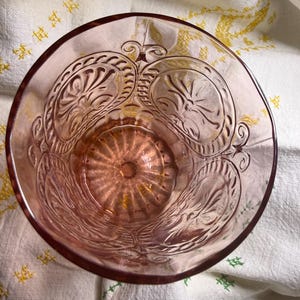 Puede incluir: Un vaso rosa vintage con intrincados patrones florales en relieve. El vaso tiene un borde ancho y un dise&ntilde;o detallado alrededor del cuenco. El vaso est&aacute; sobre un pa&ntilde;o blanco con bordados amarillos y verdes.