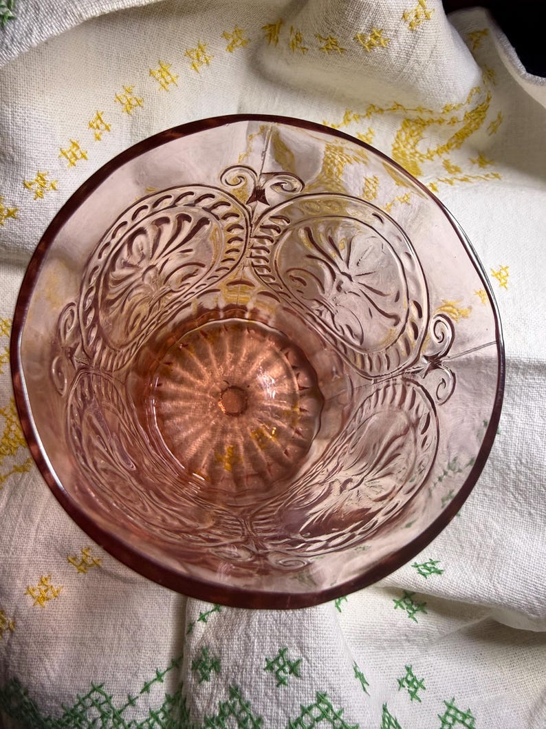Puede incluir: Un vaso rosa vintage con intrincados patrones florales en relieve. El vaso tiene un borde ancho y un dise&ntilde;o detallado alrededor del cuenco. El vaso est&aacute; sobre un pa&ntilde;o blanco con bordados amarillos y verdes.