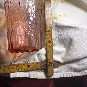 Puede incluir: Un vaso te&ntilde;ido de rosa con un dise&ntilde;o en relieve, junto a una regla de madera vintage. El vaso tiene una forma cil&iacute;ndrica alta con un patr&oacute;n detallado. La regla muestra medidas en pulgadas.