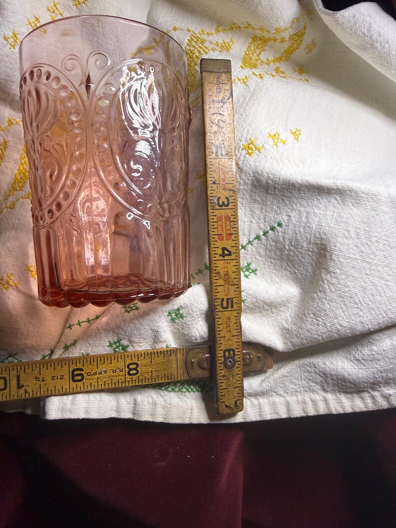 Puede incluir: Un vaso te&ntilde;ido de rosa con un dise&ntilde;o en relieve, junto a una regla de madera vintage. El vaso tiene una forma cil&iacute;ndrica alta con un patr&oacute;n detallado. La regla muestra medidas en pulgadas.