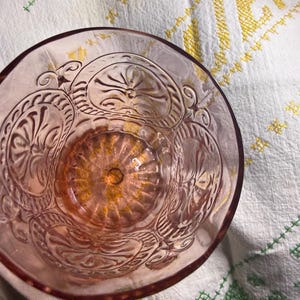 Puede incluir: Un cuenco de cristal rosa claro con un dise&ntilde;o floral en relieve ornamentado. El interior del cuenco tiene un patr&oacute;n circular texturizado. El cuenco est&aacute; sobre un pa&ntilde;o blanco con detalles bordados amarillos y verdes.