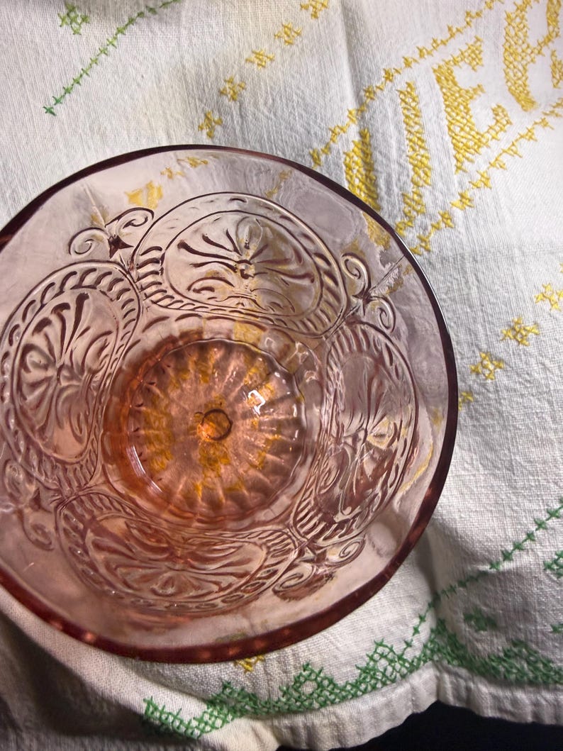 Puede incluir: Un cuenco de cristal rosa claro con un dise&ntilde;o floral en relieve ornamentado. El interior del cuenco tiene un patr&oacute;n circular texturizado. El cuenco est&aacute; sobre un pa&ntilde;o blanco con detalles bordados amarillos y verdes.