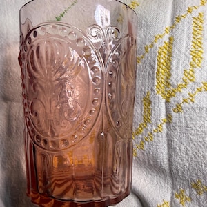 Puede incluir: Un vaso te&ntilde;ido de rosa con un dise&ntilde;o ornamental en relieve. El vaso presenta un patr&oacute;n repetitivo de formas ovaladas y motivos florales. El vaso es cil&iacute;ndrico con una parte superior ligeramente ensanchada. El fondo es un pa&ntilde;o blanco con bordados amarillos.