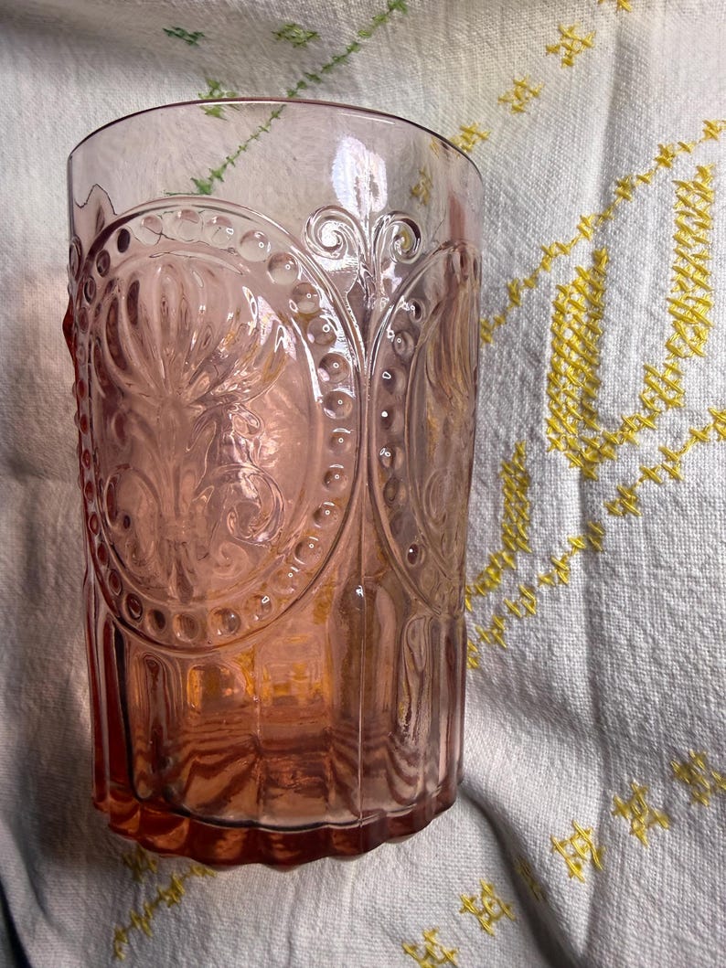 Puede incluir: Un vaso te&ntilde;ido de rosa con un dise&ntilde;o ornamental en relieve. El vaso presenta un patr&oacute;n repetitivo de formas ovaladas y motivos florales. El vaso es cil&iacute;ndrico con una parte superior ligeramente ensanchada. El fondo es un pa&ntilde;o blanco con bordados amarillos.