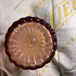 Puede incluir: Un plato de vidrio te&ntilde;ido de rosa con un borde festoneado. El plato tiene un interior texturizado y est&aacute; colocado sobre una tela blanca con detalles bordados amarillos y verdes. El vidrio parece ser antiguo.