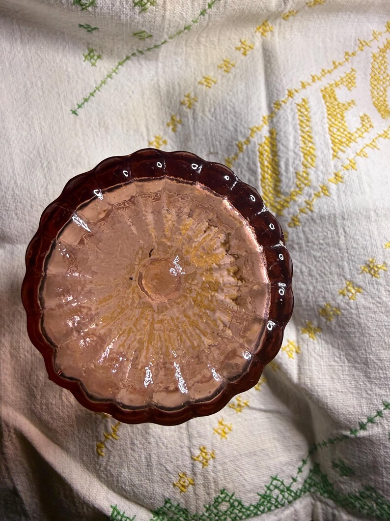 Puede incluir: Un plato de vidrio te&ntilde;ido de rosa con un borde festoneado. El plato tiene un interior texturizado y est&aacute; colocado sobre una tela blanca con detalles bordados amarillos y verdes. El vidrio parece ser antiguo.